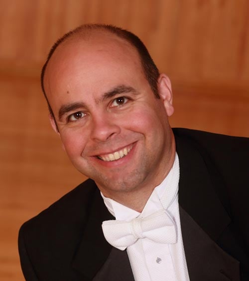 A smiling man with a bald head, dressed in a formal black tuxedo jacket and a white bow tie. The background features a warm, wood-paneled wall.