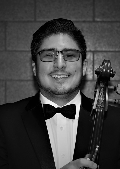 A black and white photo shows a person with short hair, glasses, and a beard, wearing a tuxedo with a bow tie, smiling while holding a cello. The background consists of a block wall.