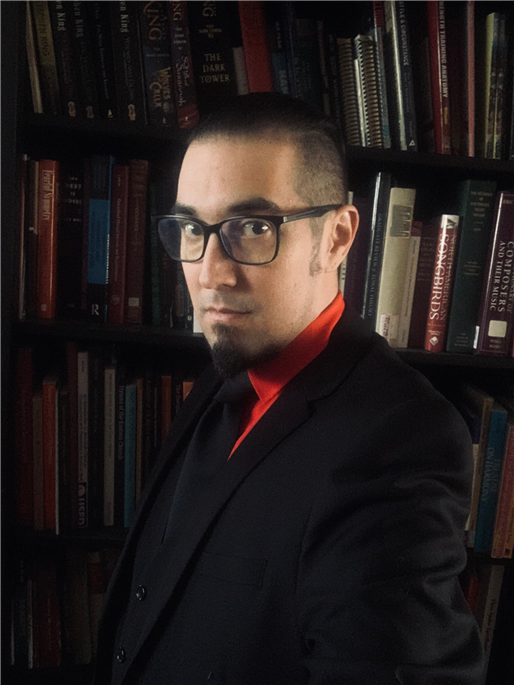 A person with short dark hair, glasses, a goatee, and wearing a black suit with a red shirt stands in front of a bookshelf filled with various books. The individual is looking directly at the camera with a serious expression.