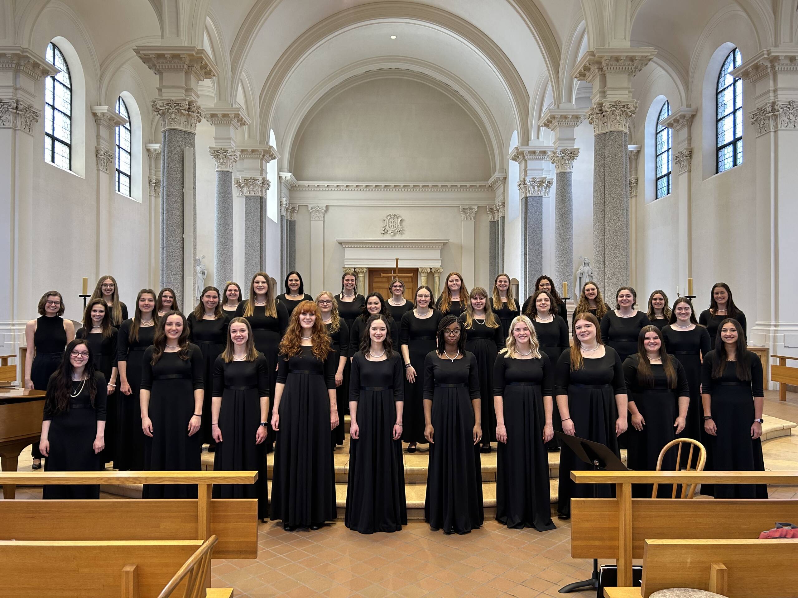 A group of women, all dressed in black gowns, stand in a semi-circular formation in a well-lit church with ornate architecture. They appear to be a choir, positioned on steps in front of an altar, and are looking forward, ready to perform.