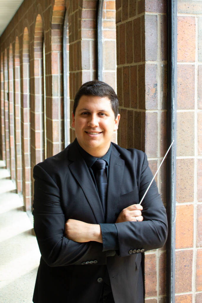 A person in a black suit and tie stands in front of a brick wall, holding a conductor's baton in their right hand and crossing their arms. They are smiling, and the background features a corridor with large windows and arches.