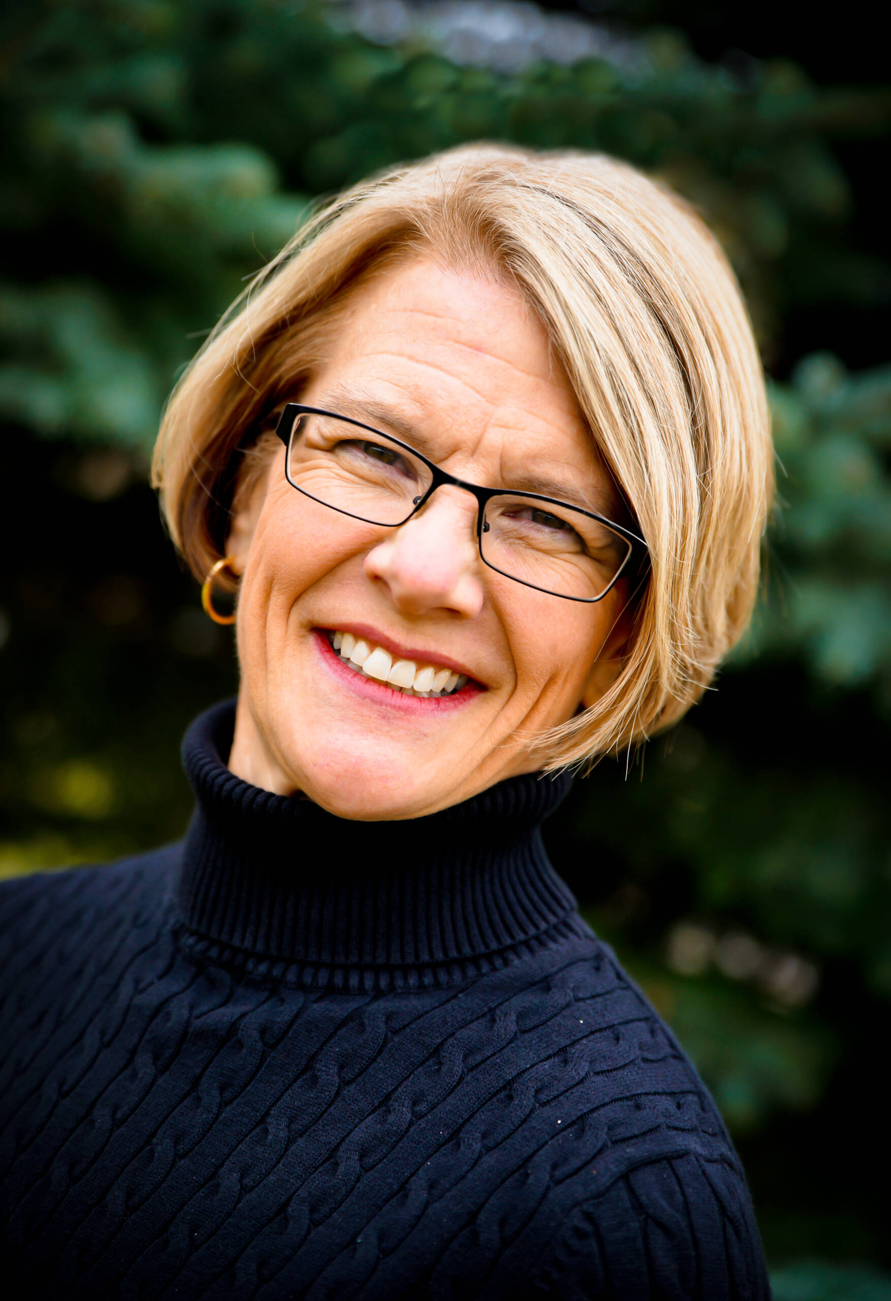 A woman with short blonde hair and glasses smiles warmly at the camera. She is wearing a black turtleneck sweater and gold hoop earrings. The background features blurred greenery.