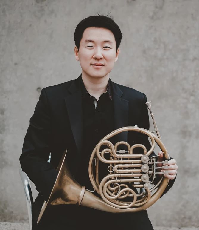 A person with short black hair is sitting against a plain wall, wearing a black suit and holding a French horn.