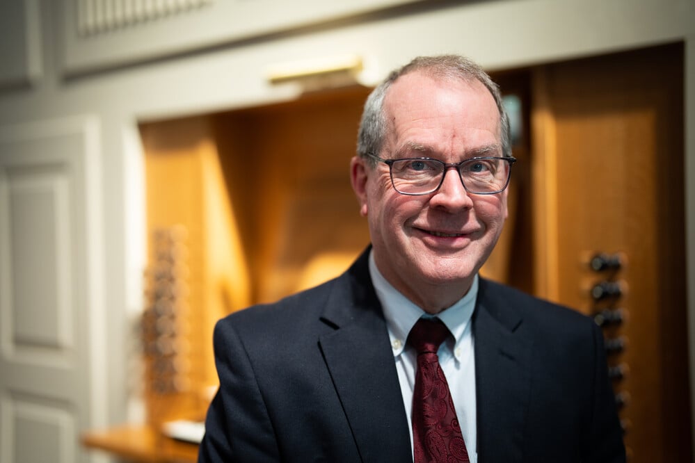 A man with glasses, wearing a dark suit and a maroon tie, smiles while standing indoors in front of a wooden structure with some visible knobs. The background appears to be blurred, highlighting the man in the foreground.