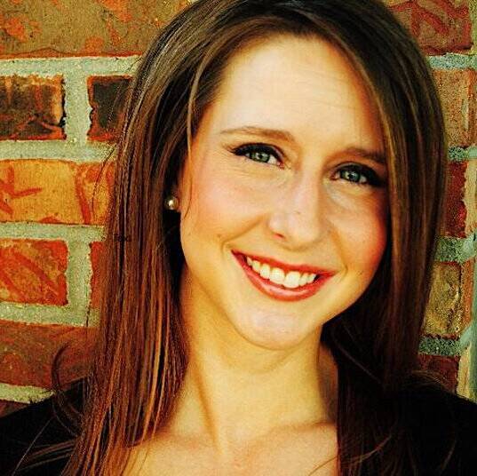 A woman with long brown hair is smiling at the camera. She has light skin, green eyes, and is wearing a black top. The background features a brick wall.