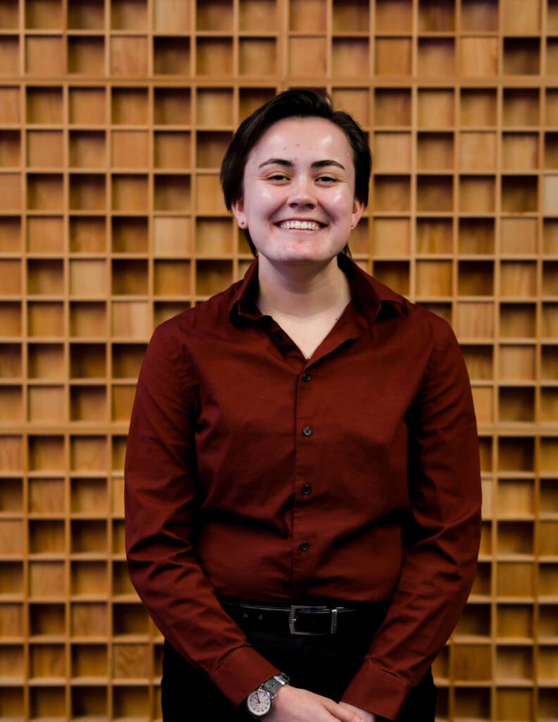 Person with short dark hair wearing a dark red button-up shirt and black pants is standing in front of a wooden grid-patterned wall, smiling warmly at the camera. They have a watch on their left wrist and their hands are clasped together in front.