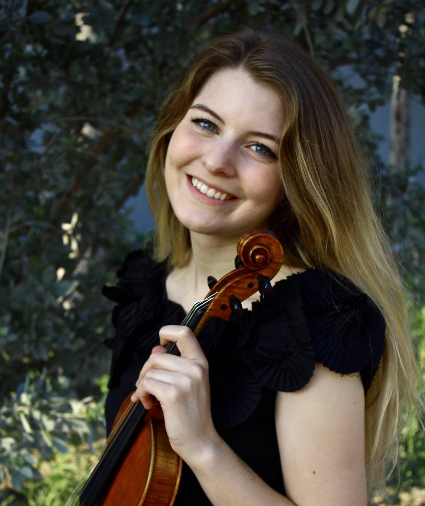 A smiling woman with long blonde hair holds a violin close to her face. She is wearing a black sleeveless top and stands in front of a background of leafy green trees.