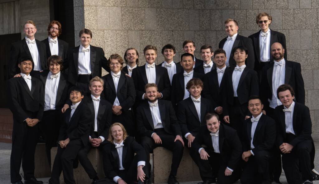 A group of twenty-four people, all dressed in formal black suits with white bow ties, pose together outdoors. Some are seated on a concrete ledge while others stand behind them, all smiling at the camera. The background features a textured wall.