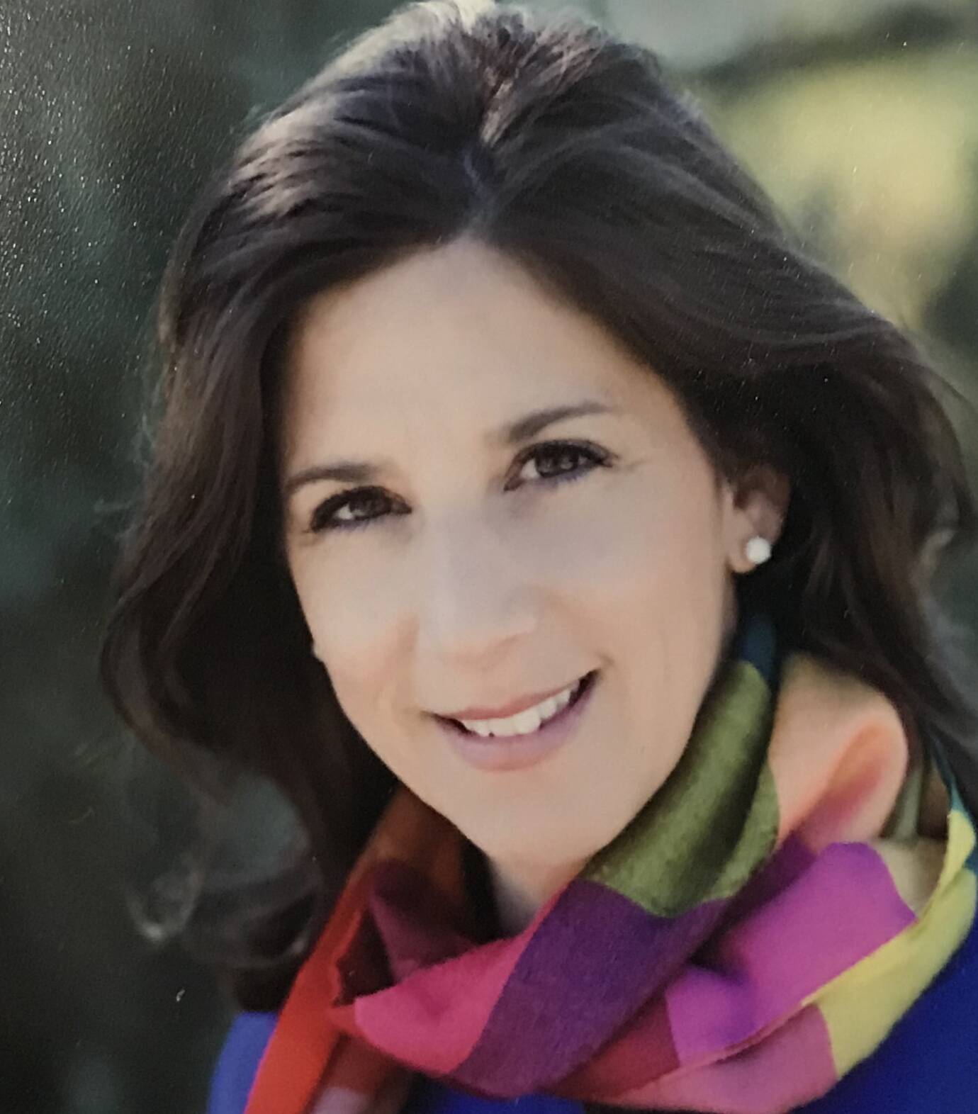 A woman with long, dark hair smiles at the camera. She is wearing a colorful scarf with shades of pink, purple, green, and yellow, along with a dark top. The background is blurred, giving a soft focus effect.