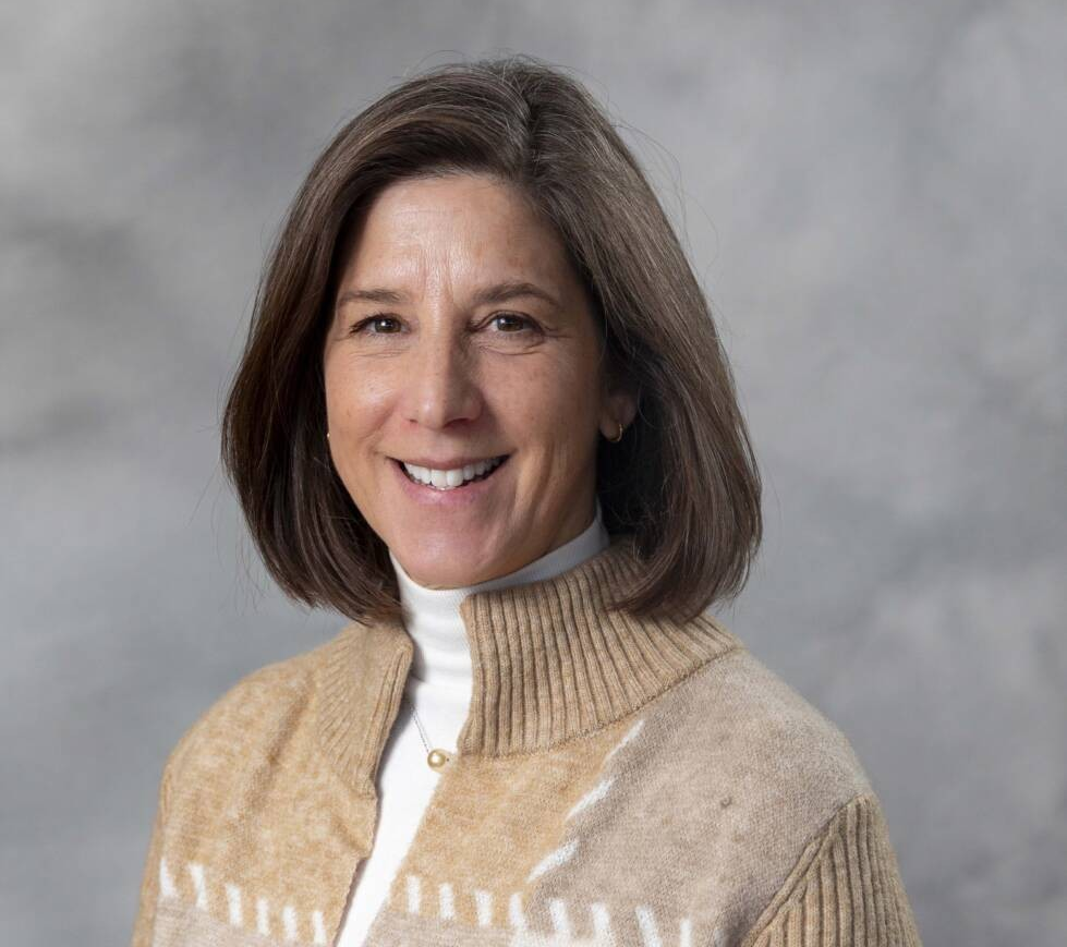 A person with shoulder-length brown hair smiles at the camera. They are wearing a beige and white sweater over a white turtleneck. The background is a textured gray.