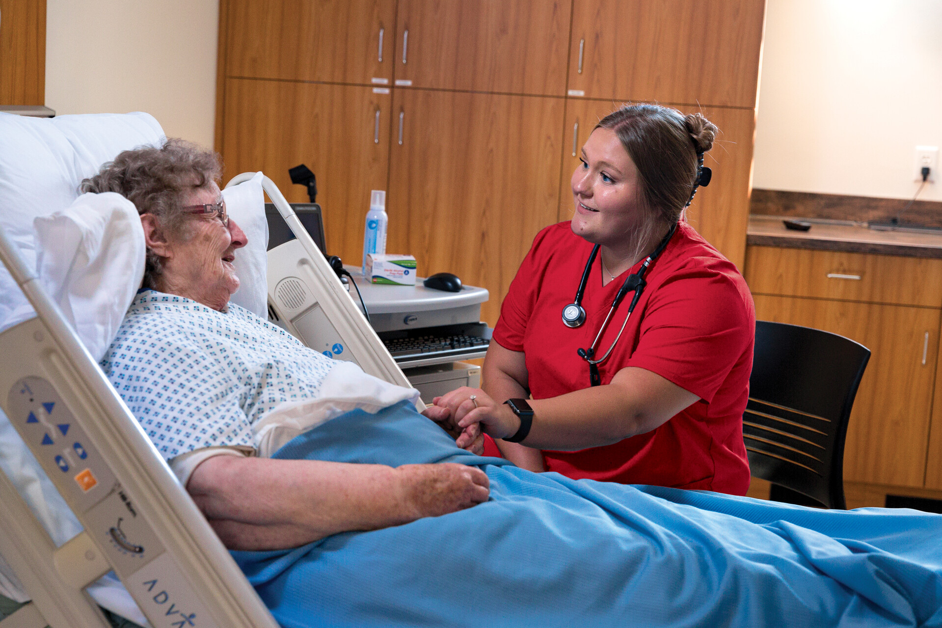 A nurse in a red uniform holds the hand of an elderly patient lying in a hospital bed. The nurse is smiling and engaging with the patient, who appears to be comfortable and attentive. The room has wooden cabinets and medical equipment nearby.