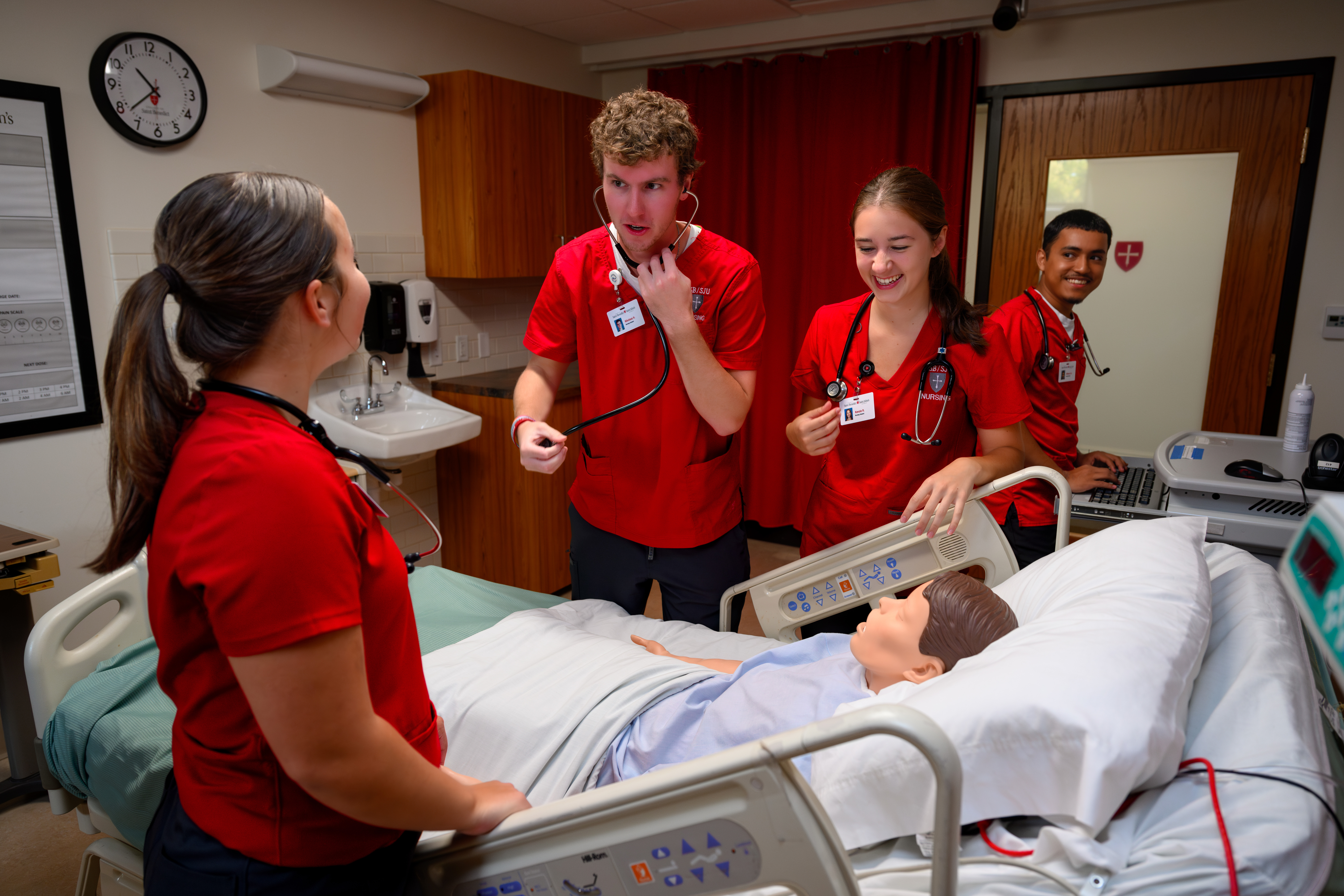 Four medical students in red scrubs practice clinical examination on a mannequin in a hospital room. They appear engaged and are using a stethoscope. A bed and medical equipment are visible in the background.