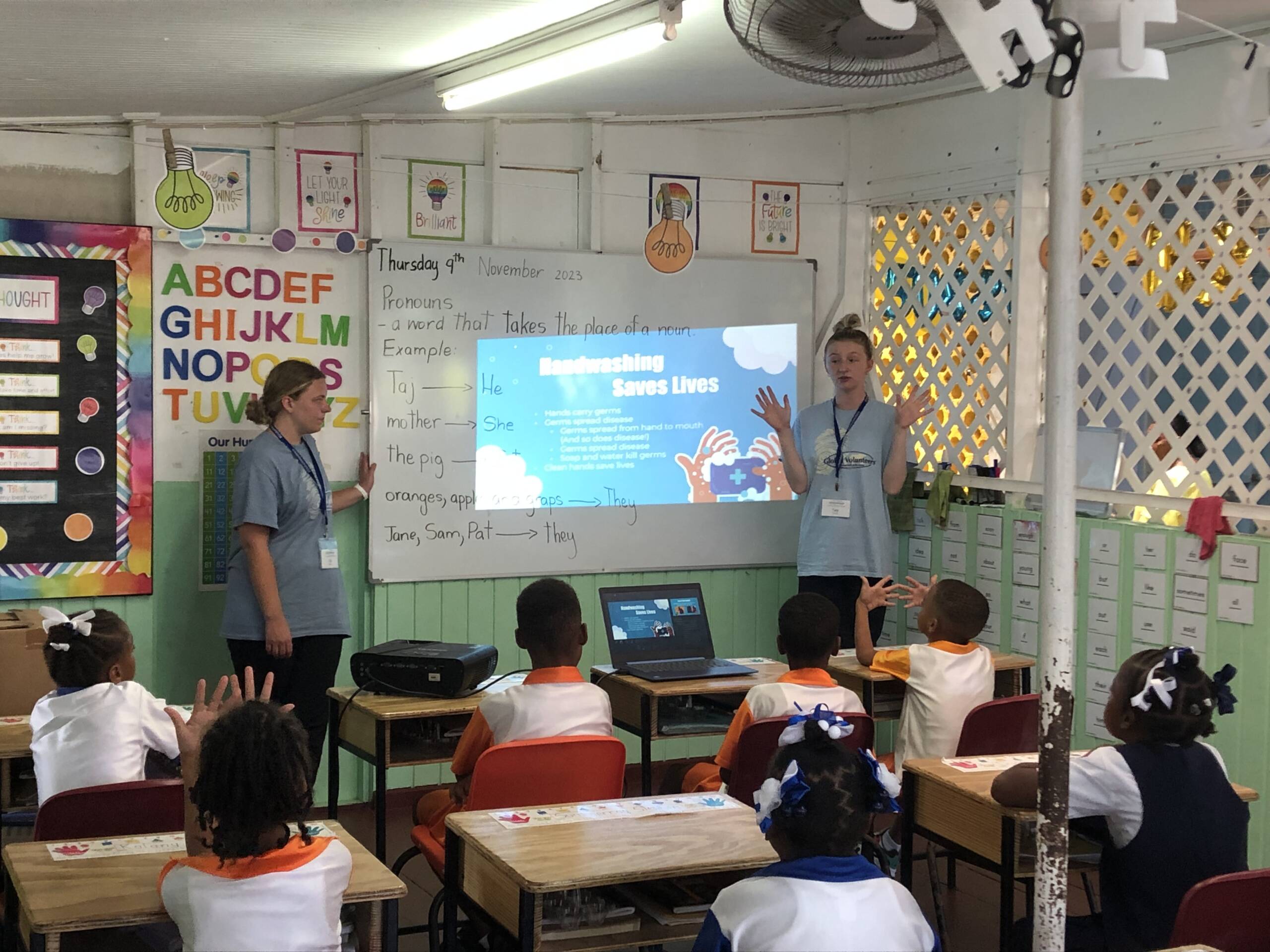 Two presenters stand in front of a classroom with young students. A slide on the screen reads "Handwashing Saves Lives." The room is decorated with educational posters and colorful alphabet charts.