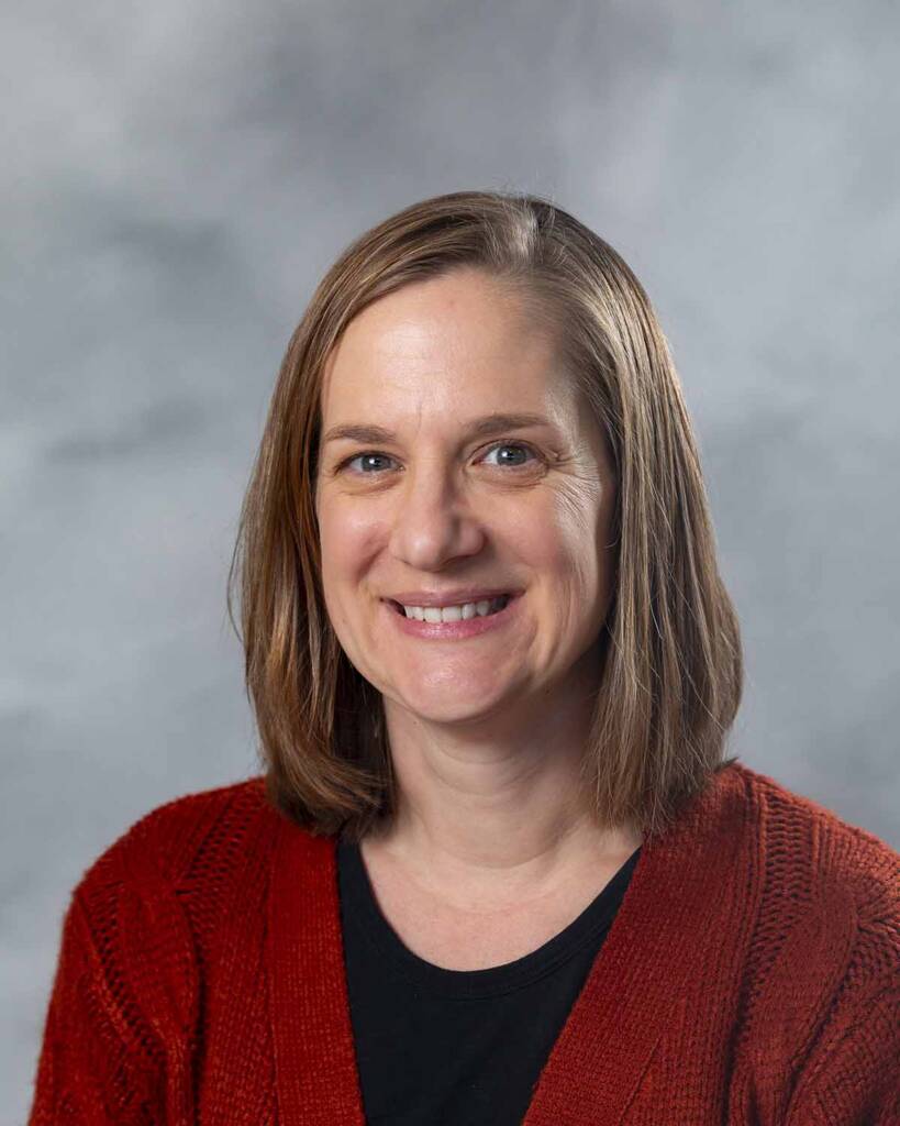 A person with shoulder-length brown hair is smiling at the camera. They are wearing a red sweater over a black top. The background is a soft, blurred gray.