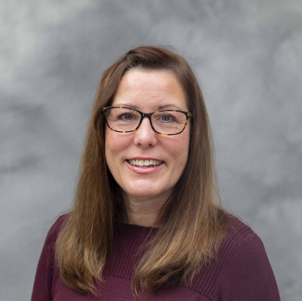 A smiling woman with long brown hair and glasses, wearing a burgundy top, stands in front of a gray background.