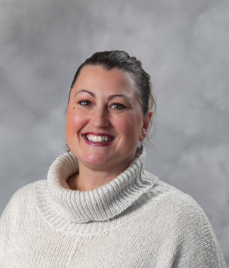 A person with dark hair pulled back is smiling widely while wearing a cozy off-white turtleneck sweater. The backdrop is a soft gray, creating a neutral and serene atmosphere.