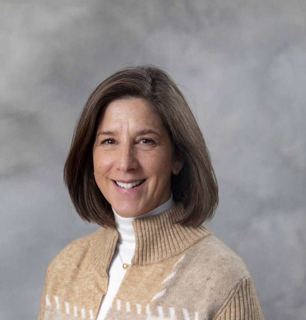 A woman with shoulder-length brown hair smiles at the camera. She is wearing a beige sweater with a white turtleneck underneath. The background is a soft gray.