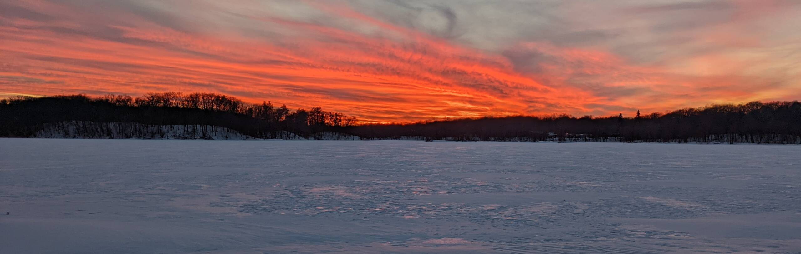 A vibrant sunset with intense orange and red hues fills the sky, casting a warm glow over a snow-covered landscape. Dark silhouettes of trees line the horizon, adding contrast to the colorful scene.