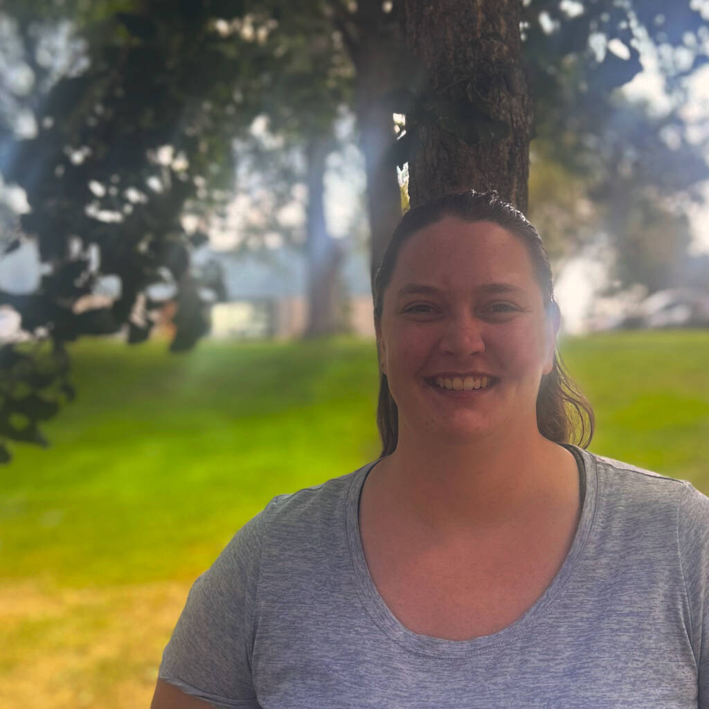 A person with long brown hair, wearing a light gray T-shirt, is standing outdoors in front of a tree. The sun is shining through the leaves, creating a soft, glowing effect. The background includes a grassy area and more trees. The person is smiling.