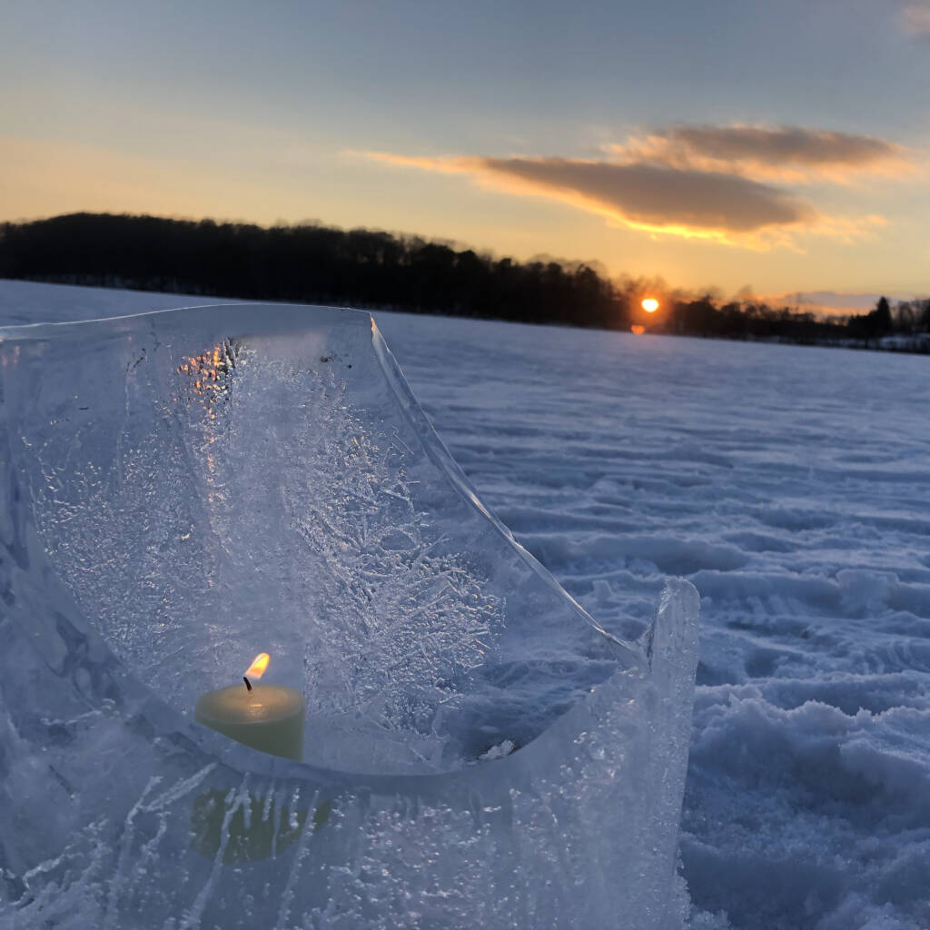 A lit candle sits inside a sculpted ice holder on a snow-covered field during sunset. The sky is tinged with orange and pink hues, and the sun is partially visible near the horizon, casting a serene glow over the landscape.