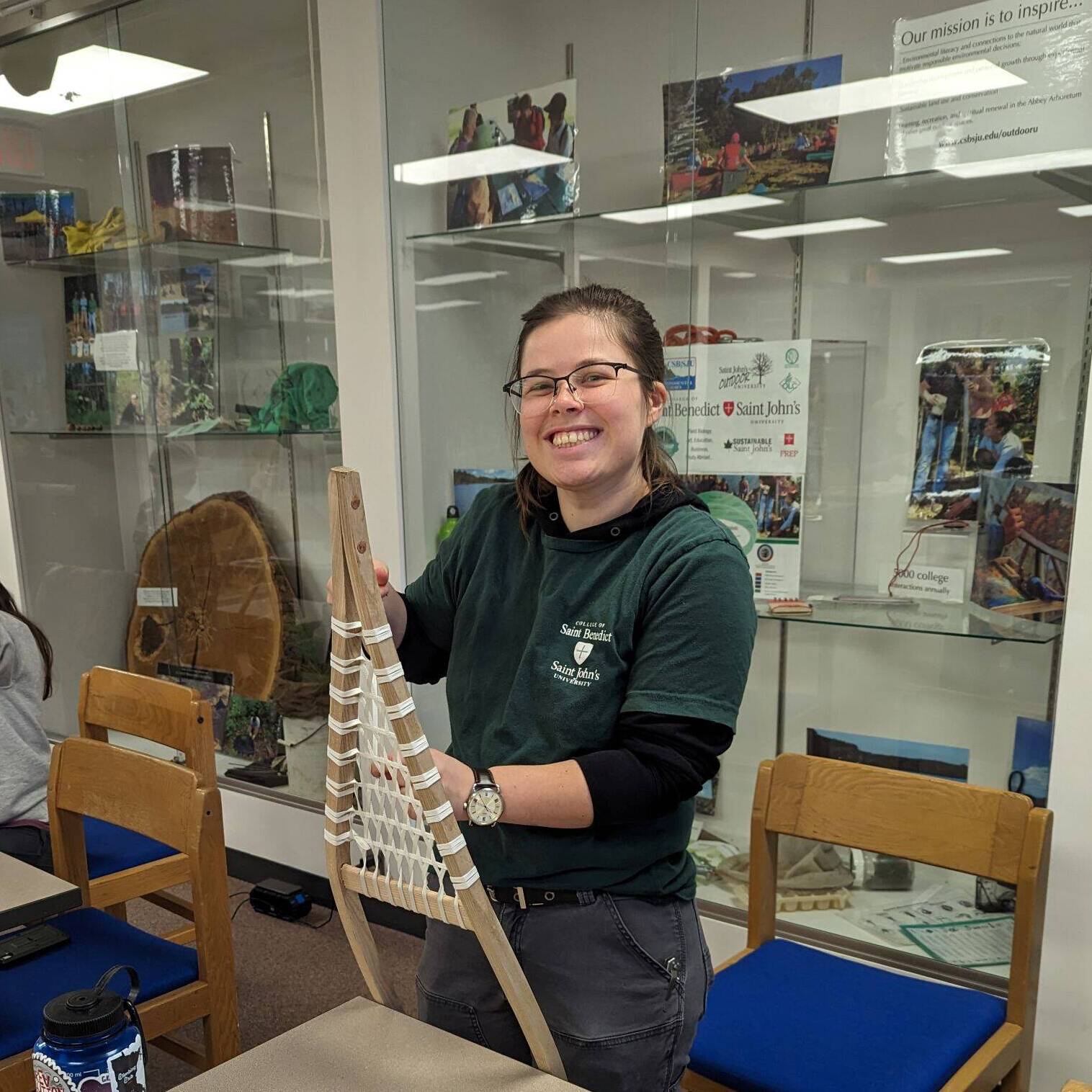 A person stands smiling at a table, holding a handmade net. The table has papers with net designs, a water bottle, and crafting supplies. Behind, shelves display various items. The setting appears to be a workshop or classroom.