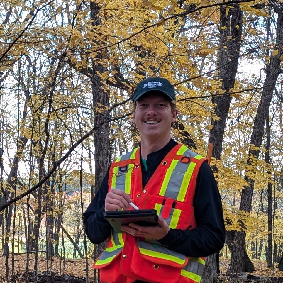 A person in a red safety vest and black cap stands in a forested area with yellow autumn leaves. They are holding a clipboard and pen, smiling at the camera. The background features tall trees and scattered foliage.