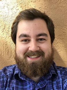 A bearded man with short brown hair is wearing a blue checkered shirt and smiling at the camera. The background features a textured beige wall.