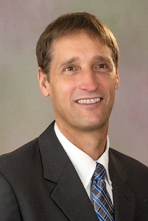 A smiling man with short brown hair, wearing a dark suit, white shirt, and striped tie, poses for a professional photo against a neutral background.