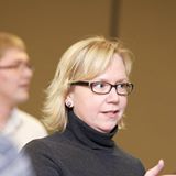 A woman with short blonde hair and glasses is wearing a black turtleneck shirt. She is indoors and her mouth is slightly open as if she is speaking. Another person, out of focus, is in the background wearing a white shirt.