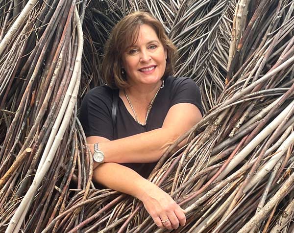 A woman with short brown hair and a black short-sleeved top is smiling and leaning with her arms resting on intertwined woven branches in the background. She is wearing a silver watch and a necklace.