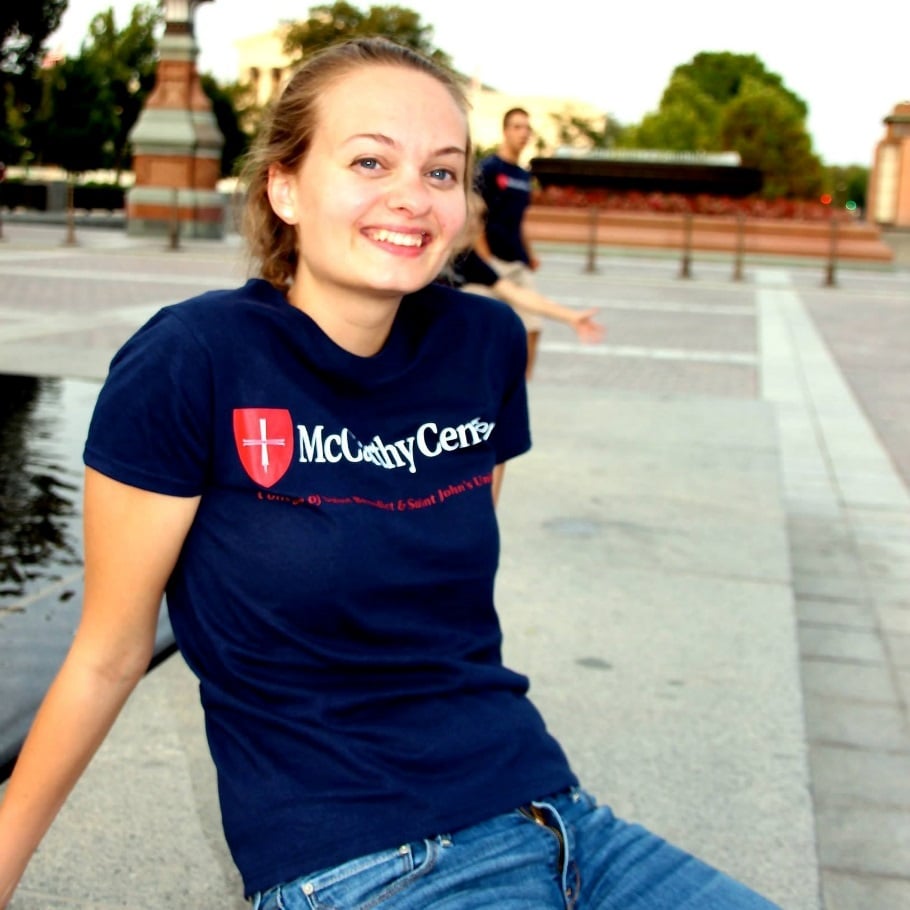 A young woman with light brown hair tied back is sitting on a ledge, smiling at the camera. She is wearing a dark blue T-shirt with the words "McGinty Center" printed in white and blue jeans. There is another person blurred in the background along with trees and a building.