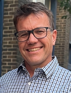 A man with short brown hair and glasses smiles while wearing a blue and white checkered shirt. The background features a brick wall and some foliage.