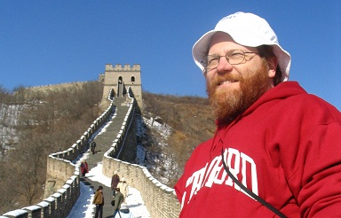 A man with a beard, wearing glasses, a white hat, and a red hoodie, stands smiling in front of the Great Wall of China. Several people can be seen walking along the wall, with clear blue skies above and a lightly snow-dusted landscape surrounding the wall.