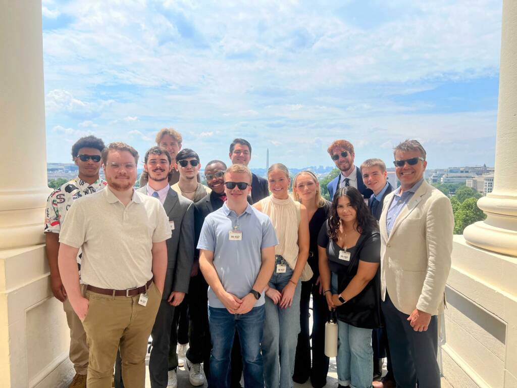 A group of people smiling and posing on a balcony with an expansive city skyline in the background. They are dressed in a mix of casual and business attire, and the sky is partly cloudy.