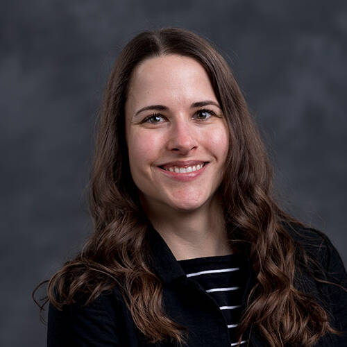 A woman with long, wavy brown hair smiles at the camera against a gray, textured background. She is wearing a black jacket over a black and white striped top.