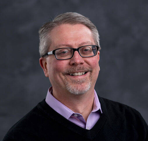 A man with short gray hair, glasses, and a neatly trimmed beard smiles warmly at the camera. He is wearing a black sweater over a light purple collared shirt. The background is a muted dark gray, creating a professional and approachable atmosphere.