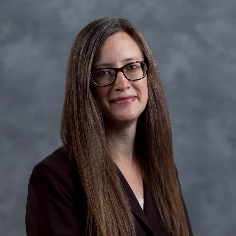 A woman with long, straight brown hair and glasses wearing a dark blazer poses against a gray background.