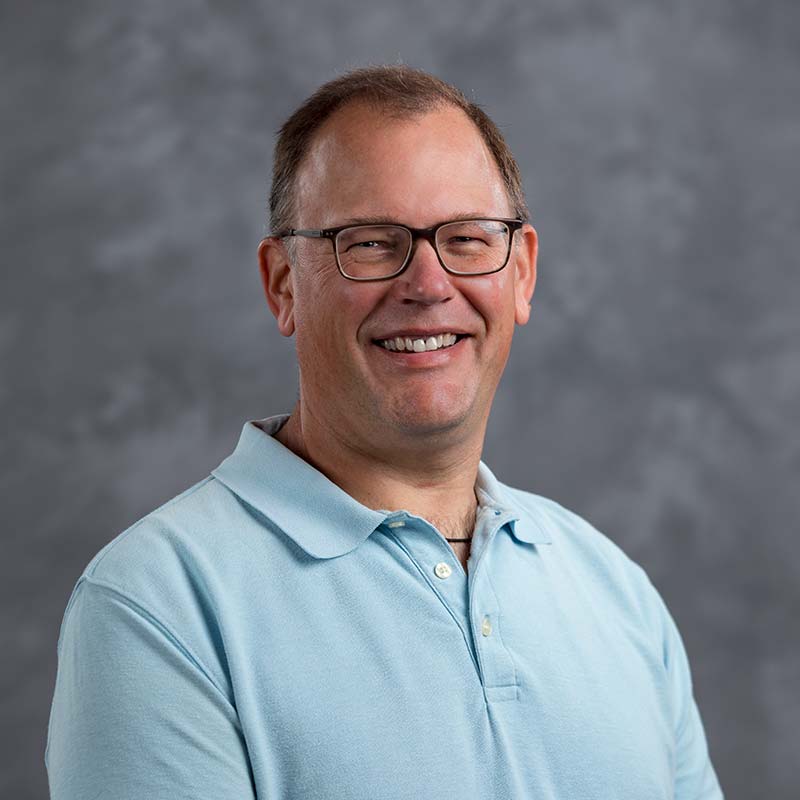 A smiling man wearing glasses and a light blue polo shirt stands in front of a grey, textured background.