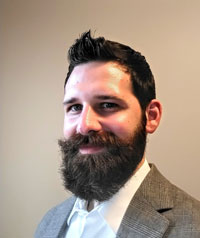 A man with dark hair and a full beard smiling. He is wearing a gray suit jacket over a white dress shirt. The background is a plain, light-colored wall.