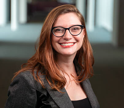 A woman with long, auburn hair and glasses smiles while wearing a professional gray blazer over a black top. She stands in a softly lit hallway with blurred background, projecting a professional and approachable demeanor.