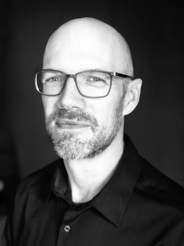 A black and white portrait of a bald man with glasses and a beard. He is facing slightly to the left and looking directly at the camera with a slight smile. The background is dark, creating a contrast with his light skin tone.