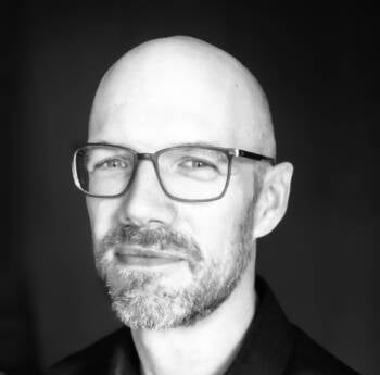 A black and white portrait of a bald man with glasses and a beard. He is facing slightly to the left and looking directly at the camera with a slight smile. The background is dark, creating a contrast with his light skin tone.