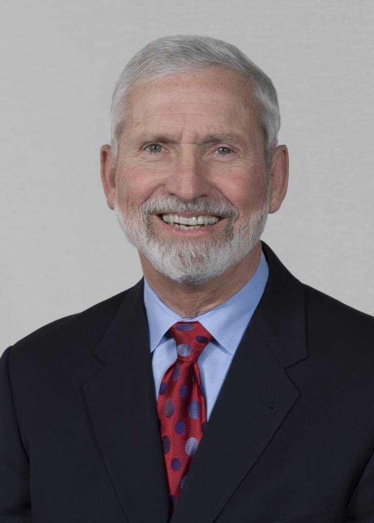 A smiling older man with white hair and a beard, wearing a black suit, blue shirt, and red tie with purple dots, poses against a plain background.