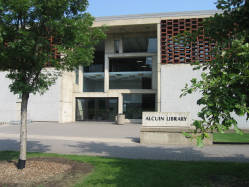 Exterior view of a modern library building with large windows and a sign that reads "Alcuin Library." Surrounded by trees and a clear pathway.