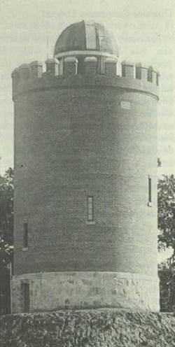 A black and white photo of a tall, cylindrical brick tower with a dome roof and battlement-style parapet. The structure is set on a stone base, surrounded by trees.