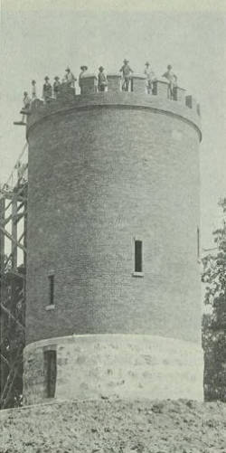 A vintage photograph of a tall, cylindrical brick tower with a crenellated parapet. Several people are standing along the top edge. The lower part of the structure is made of stone, and there is scaffolding visible on the left side.