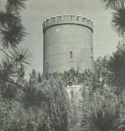 A black-and-white image of a cylindrical stone tower with crenellations at the top, surrounded by dense trees and foliage. The perspective is slightly upward, highlighting the tower's prominence against the sky.