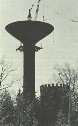 A black and white image of a tall water tower and a shorter stone building with a crenellated roof, surrounded by bare trees. A crane is visible next to the water tower against the cloudy sky.