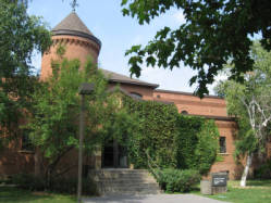 A small, red brick building with a round tower topped by a conical roof. The structure is partially covered with green ivy. It is surrounded by lush green trees and bushes, and a pathway leads to its entrance.