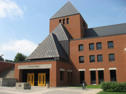 A red-brick building with a sloped, gray metal roof and an entrance labeled "Student Commons." The structure features modern geometric design elements, with windows and a small tower. A bicycle is parked outside under a clear blue sky.